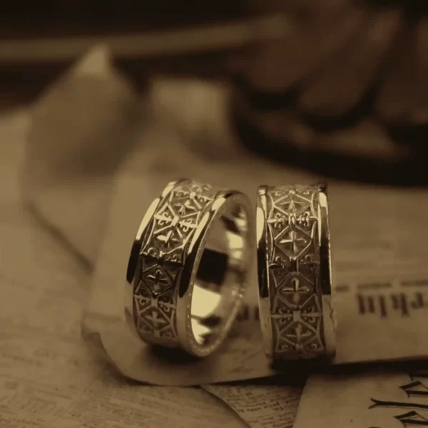 In the image, a pair of matching rings crafted from light silver is showcased, exuding an aura of elegance and sophistication. These rings, from the renowned Silveringdom, feature intricate designs reminiscent of a knight’s regalia, symbolizing strength and nobility. Each ring is adorned with a delicate pattern that resembles an iris, adding a touch of natural beauty and uniqueness. The craftsmanship highlights the meticulous attention to detail, making these rings a perfect symbol of unity and commitment. The background, with its subtle, blurred texture, enhances the timeless appeal of these exquisite pieces, making them a standout choice for those seeking both tradition and modernity in their jewelry.