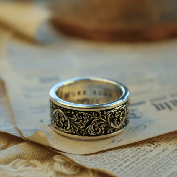 This exquisite ring from Silveringdom features intricate scrollwork that showcases the artistry and craftsmanship of its design. The scrollwork is in high relief, creating a three-dimensional effect that catches the light beautifully. The ring’s polished silver surface contrasts elegantly with the detailed scrollwork, adding depth and texture to the piece. Resting on an aged piece of paper, the ring exudes a sense of antiquity and timeless elegance. The combination of the ornate metalwork and the historical backdrop suggests a story of heritage and tradition, making this ring not just a piece of jewelry, but a work of art that carries a rich narrative.