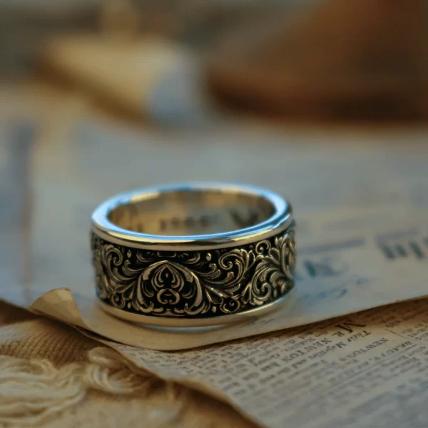 This pair of matching rings from Silveringdom is crafted from dark silver, featuring intricate scrollwork in high relief. The ornate patterns stand out against the polished surface, creating a stunning three-dimensional effect. The rings rest on an aged piece of parchment with handwritten text, enhancing their antique and timeless appeal. The detailed scrollwork and the dark silver material give these rings a regal and majestic quality, making them perfect for those who appreciate fine craftsmanship and historical elegance. The combination of the ornate metalwork and the historical backdrop suggests a story of heritage and tradition, making these matching rings not just pieces of jewelry, but works of art that carry a rich narrative.