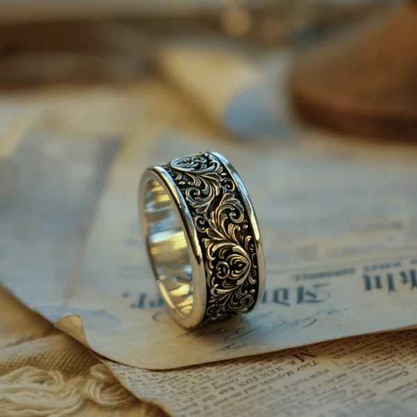 This pair of matching rings from Silveringdom, crafted from dark silver, features intricate scrollwork in high relief. The ornate patterns create a stunning three-dimensional effect, standing out against the polished surface. Resting on an aged piece of parchment with handwritten text, these rings exude an antique and timeless charm. The detailed scrollwork and dark silver material give the rings a regal and majestic quality, perfect for those who appreciate fine craftsmanship and historical elegance. The combination of the ornate metalwork and the historical backdrop suggests a story of heritage and tradition, making these matching rings not just pieces of jewelry, but works of art that carry a rich narrative.