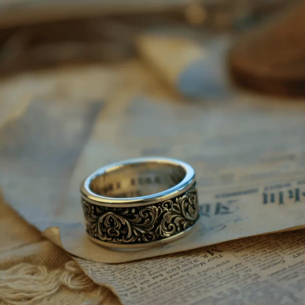 This pair of matching rings from Silveringdom is crafted from dark silver, featuring intricate scrollwork in high relief. The detailed patterns create a striking three-dimensional effect, standing out against the smooth, polished surface. Placed on an aged piece of parchment with handwritten text, these rings exude a sense of timeless elegance and historical charm. The dark silver material and ornate scrollwork give the rings a regal and sophisticated appearance, perfect for those who appreciate fine craftsmanship and vintage aesthetics. The combination of the intricate metalwork and the antique backdrop suggests a rich narrative of heritage and tradition, making these matching rings not just pieces of jewelry, but true works of art.