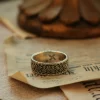In the heart of Silveringdom, a vintage vow is beautifully captured in this image of exquisite wedding rings. The ring, crafted from dark silver, showcases the meticulous art of handcraft. Its intricate design speaks to a gender-neutral aesthetic, making it a perfect symbol for any couple. The band is adorned with delicate patterns, reflecting the timeless elegance and attention to detail that Silveringdom is renowned for. Each ring is a bespoke creation, allowing couples to customize their symbols of love to reflect their unique bond. This piece harmonizes tradition with modernity, embodying a perfect blend of strength and delicacy. Resting on an aged page, the ring tells a story of love that transcends time, making it a cherished heirloom for generations to come.