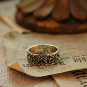 In the heart of Silveringdom, a vintage vow is beautifully captured in this image of exquisite wedding rings. The ring, crafted from dark silver, showcases the meticulous art of handcraft. Its intricate design speaks to a gender-neutral aesthetic, making it a perfect symbol for any couple. The band is adorned with delicate patterns, reflecting the timeless elegance and attention to detail that Silveringdom is renowned for. Each ring is a bespoke creation, allowing couples to customize their symbols of love to reflect their unique bond. This piece harmonizes tradition with modernity, embodying a perfect blend of strength and delicacy. Resting on an aged page, the ring tells a story of love that transcends time, making it a cherished heirloom for generations to come.
