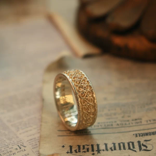 The image showcases a stunning wedding ring from Silveringdom, embodying the essence of a vintage vow. This handcrafted piece, made from light silver, exudes timeless elegance and meticulous craftsmanship. The intricate detailing on the ring highlights the brand’s dedication to creating unique, customizable jewelry. Designed to be gender-neutral, this wedding ring appeals to a wide range of tastes and preferences, making it a perfect symbol of love and commitment for any couple. The ability to customize the ring ensures that each piece is as unique as the love story it represents. Silveringdom’s commitment to quality and individuality shines through in this exquisite example of their work.
