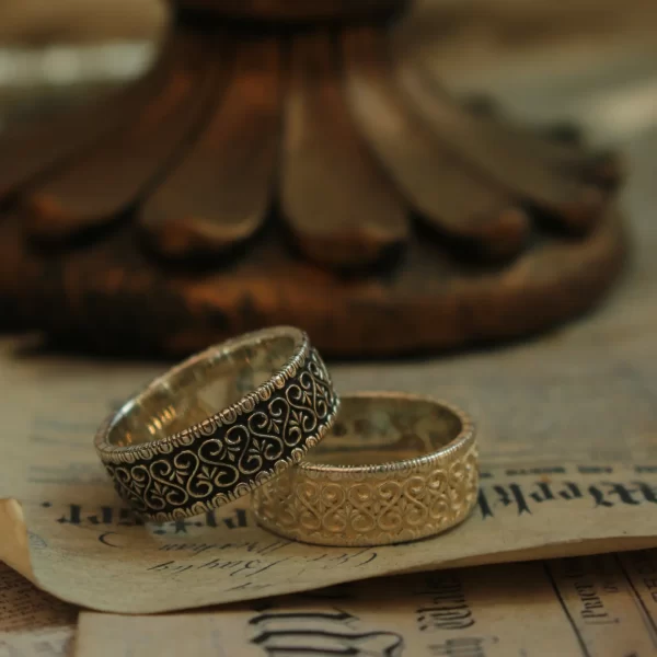 In the heart of Silveringdom, a pair of matching rings rests elegantly on a vintage book, symbolizing a timeless vintage vow. These rings are a testament to exquisite handcraft, showcasing the meticulous artistry involved in their creation. One ring features a light silver finish, capturing the essence of simplicity and elegance, while its counterpart boasts a dark silver hue, adding a touch of mystery and sophistication. The androgynous design of these rings makes them perfect for any couple, transcending traditional gender norms. Each piece is customized to reflect the unique bond between two individuals, making them not just accessories, but meaningful symbols of love and commitment. The delicate interplay of light and shadow on the rings highlights their intricate details, celebrating both unity and individuality. These rings from Silveringdom are more than just jewelry; they are handcrafted treasures that tell a story of enduring love.