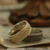 In the heart of Silveringdom, a pair of matching rings rests on an aged parchment, embodying the essence of a vintage vow. These rings are the result of meticulous handcraft, shaped by skilled artisans who understand the beauty of uniqueness. The larger ring features an intricate pattern etched into light silver, catching the eye with its delicate play of light and shadow. Its companion, slightly smaller, is adorned with a similar design but stands out in dark silver, offering a bold contrast that speaks to an androgynous appeal—neither overtly masculine nor feminine but transcending traditional boundaries. Both rings invite customization, ensuring that no two pairs are identical; each set tells its own story. This image captures more than just jewelry; it represents a commitment to craft and individuality—a testament to love’s diverse expressions.