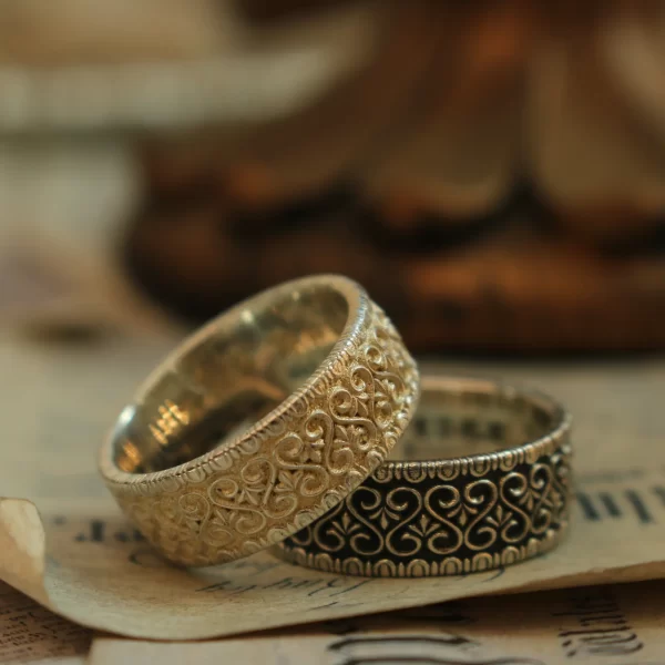 In the heart of Silveringdom, a pair of matching rings rests on an aged parchment, embodying the essence of a vintage vow. These rings are the result of meticulous handcraft, shaped by skilled artisans who understand the beauty of uniqueness. The larger ring features an intricate pattern etched into light silver, catching the eye with its delicate play of light and shadow. Its companion, slightly smaller, is adorned with a similar design but stands out in dark silver, offering a bold contrast that speaks to an androgynous appeal—neither overtly masculine nor feminine but transcending traditional boundaries. Both rings invite customization, ensuring that no two pairs are identical; each set tells its own story. This image captures more than just jewelry; it represents a commitment to craft and individuality—a testament to love’s diverse expressions.