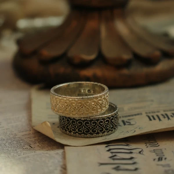 In the image, a pair of wedding rings from Silveringdom’s vintage vow collection is beautifully displayed. These rings showcase exquisite handcraft, with intricate patterns etched into their bands. One ring features a light silver hue, offering a subtle contrast to the other’s dark silver tone. Both rings embody an androgynous design, appealing to any individual regardless of gender. The craftsmanship suggests that these rings can be customized to suit personal tastes while maintaining their timeless elegance. The background includes an aged piece of paper with printed text, enhancing the vintage atmosphere surrounding these symbols of eternal commitment. This image captures not only the beauty of bespoke jewelry but also evokes a sense of history and tradition through its composition and props used, aligning perfectly with the concept of a lifelong vow made on one’s wedding day.