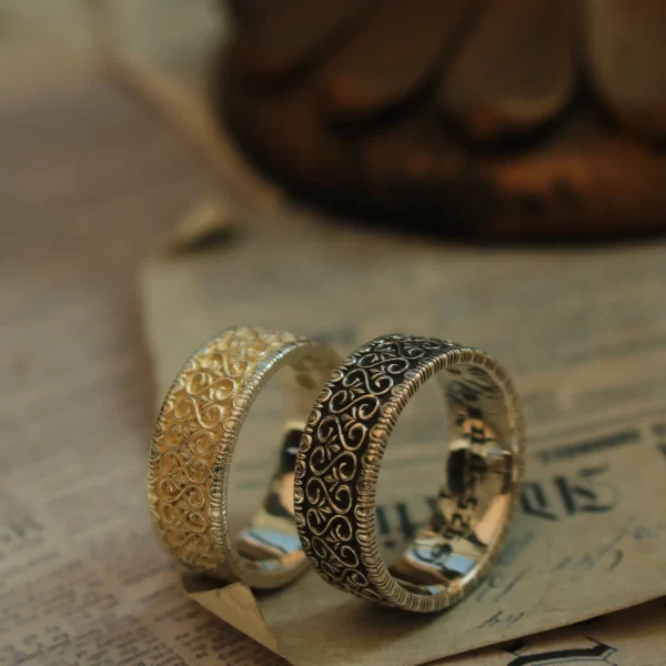 In the image, two beautifully crafted wedding rings are showcased, embodying the essence of ‘silveringdom.’ The ring on the left, with its light silver hue, features intricate patterns that evoke a sense of a vintage vow, highlighting the artistry of handcraft. Its counterpart, in dark silver, boasts a unique design that speaks to the meticulous customization involved. Both rings are gender-neutral, making them perfect for any couple seeking a timeless symbol of their union. The contrast between the light silver and dark silver rings not only accentuates their individual beauty but also complements them as a harmonious pair. These wedding rings are a testament to the blend of traditional craftsmanship and modern design, offering a personalized touch that caters to diverse tastes and preferences.